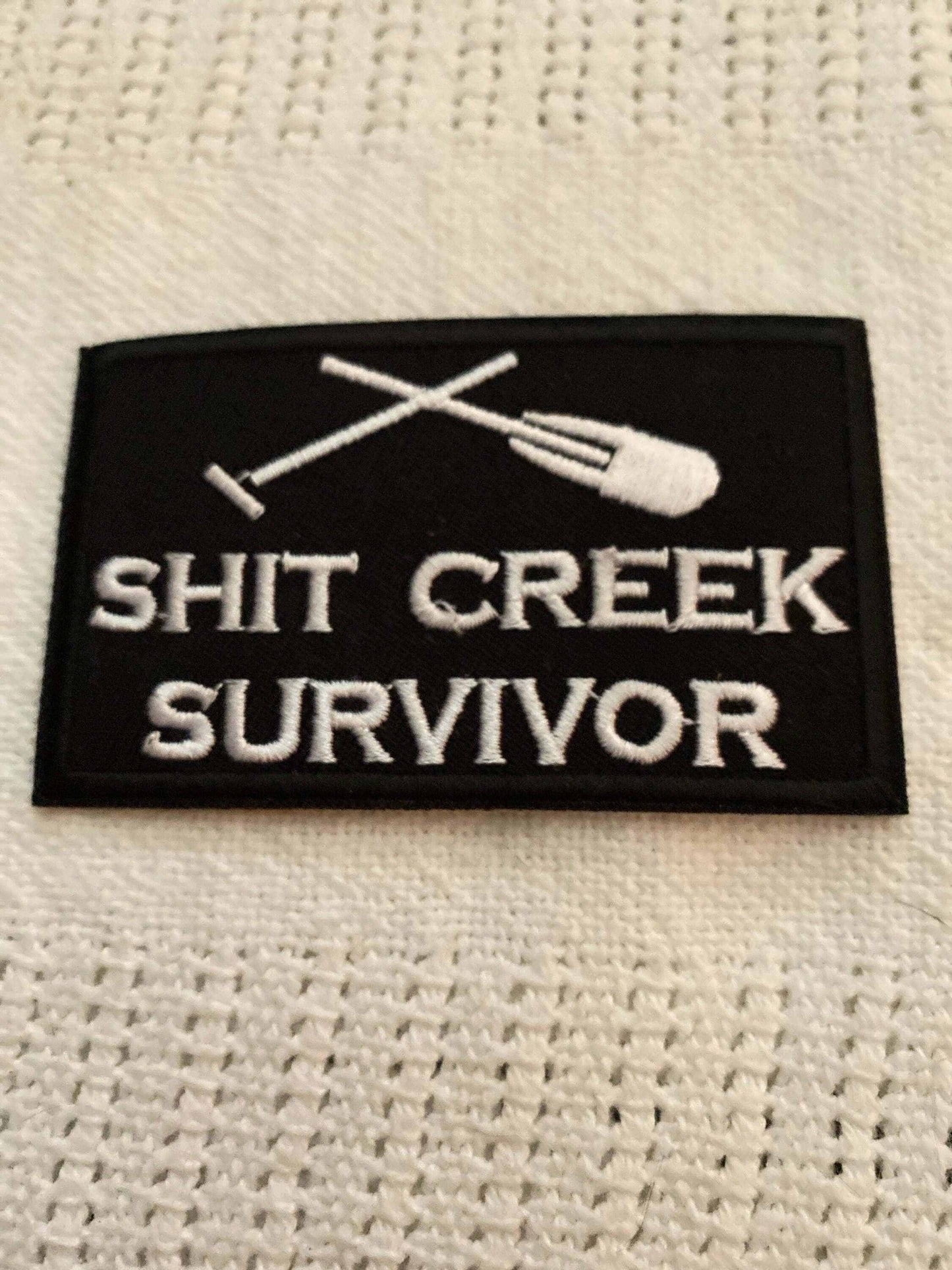 Black and white embroidered patch with crossed paddle design and text "Shit Creek Survivor" representing Creek Survivor outdoor gear brand.